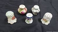 Set of five vintage porcelain salt and pepper shakers, floral patterns, all missing bottom plugs, arranged on black fabric.