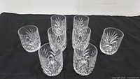 Group photo of 8 clear crystal on the rocks liquor glasses arranged on black fabric showing their general shape and design.