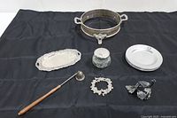 Overall lot including silver serving stand, tray, vanity jar, plates, candle snuffer, pewter lid, and napkin ring arranged on black fabric.