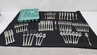 Image showing multiple flatware pieces laid out on a black cloth, including knives, forks, spoons, and smaller forks in different styles and finishes.