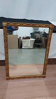 Front view of the rectangular gold gilded mirror framed with detailed carved designs, displayed against a warehouse background.