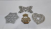 All four trivets arranged on a white surface. Top left is silver six-point star, top center is silver butterfly, bottom left is gold lotus flower, bottom right is silver heart.