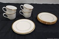 Four white porcelain cups with gold trim stacked in pairs, shown with several white and gold dessert plates aligned next to them.