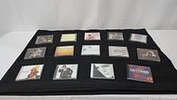 15 music CDs laid out in a grid on black cloth, front covers visible showing various artists and album artwork.