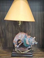 Full view of the table lamp showing the Asian war horse base and beige shade on wooden surface with dark wood panel background.