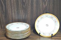 Stack of 8 antique floral porcelain plates with gold trim and 1 plate displayed upright on stand