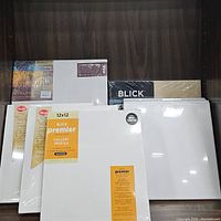 Photo showing multiple wrapped white canvases including four 12x12 Blick Premier canvases and unmarked canvases behind.