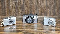 Three Canon cameras side by side on a wooden surface: two smaller silver models and one larger gray model in the center.
