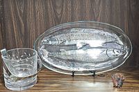 Photo showing all three items together on wood surface against wood paneled background: oval fish platter, crystal ice bucket with metal tongs inside, and multicolored glass sake cup.
