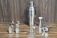 Full set including cocktail shaker, double-ended jigger, drinking straws, muddler, cap strainer, and six metal shot glasses displayed on wood surface.