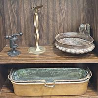 Four metal items displayed on wood shelf: cast iron nutcracker, twisted brass bracket, fluted metal bowl, rectangular copper pot.
