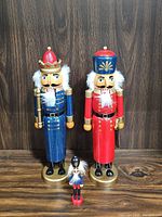 Three wooden nutcracker figurines shown against wooden background: two large soldiers in blue and red, one small figurine