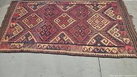 Full view of vintage tribal rug showing entire rectangular shape and multicolor geometric diamond motifs in red, cream, brown, and blue colors on concrete floor.