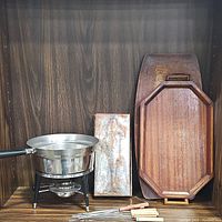 Vintage kitchen items displayed on wood background including fondue pot on stand, rectangular wood serving tray, wooden cutting board, metal fish mold, and four wooden fondue forks.