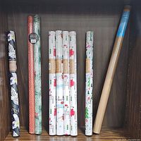Nine rolls of wrapping paper neatly arranged on a wooden shelf. Seven rolls have white background decorated with Santa Claus and Christmas trees, one roll is red with white dots, one roll has floral pattern, and one kraft paper roll is visible.