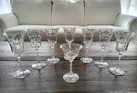 Group photo showing 6 crystal cut wine glasses and one smaller parfait dish, all with etched leaf patterns and ornamental stems, displayed on a wood table with couch backdrop.