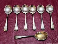 Set of 6 Birks Regency Plate soup spoons with 1 smaller teaspoon displayed on burgundy fabric, all with monogram 'G' on handles.