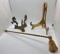 Three vintage brass items: folding easel, decorative tripod stand, and twisted candle wick snuffer lying flat.