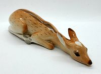 Porcelain baby fawn figurine lying down, side angle showing detailed face and front legs folded beneath body, glossy finish with natural fawn colors.