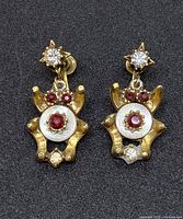 Close-up of vintage gold-tone Art Deco style dangle earrings showing ruby-colored stones, clear rhinestones, mother of pearl center and screwback closure, with some visible tarnish and wear.