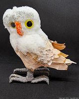 Front-right angled view of the handcrafted snowy owl figurine showing the white textured fabric covering the head and body, orange beak, large yellow-eyed features, and birch bark wings and tail with detailed grey feet.