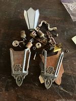 View of all parts including two metal sconce backplates with vintage decorative patterns, one geometric white glass shade, bulb sockets, nuts, screws and other metal parts scattered on a wood surface.