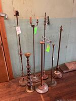 Photo showing seven antique floor lamps and a large brass candlestick clustered against a wall, various designs visible including two candle-style bulb holders and turned brass candlestick.