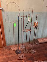 Six antique floor lamp and stand bases of various designs including Art Deco smoking stand and birdcage stand against a wall.