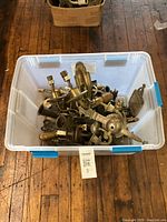 Plastic bin containing various brass and metal wall light fixtures, parts and bases, shown on wooden floor.