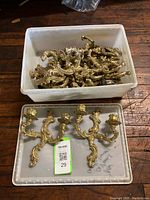 Overhead view of multiple ornate brass sconces in a plastic container and loose on lid, showing 14 total pieces with detailed scrollwork design.