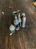 Photo showing two bronze lamp bases with detailed openwork designs, two smooth bronze lamp covers, and a green bug zapper unit on a wooden floor.