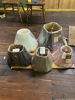 Five new discontinued lampshades arranged together on floor, some in plastic wrapping with attached labels showing measurements