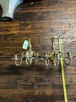 Photo of two antique brass colonial wall sconces laid flat on wooden floor with measuring tape showing approximate length and tag number attached.