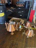 Overview of nine wood and copper table lamps with shades and cords arranged on wooden floor