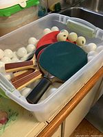 Plastic tote containing multiple white ping pong balls and some ping pong paddles inside.