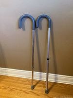Two adjustable walking canes with blue curved handles, leaning against a wall with new gray rubber tips on wooden floor.