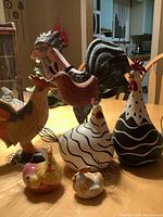 Photo of seven decorative chicken figurines including wooden, glass, metal, and ceramic types on a wooden table with kitchen background.