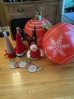 Two large red inflatable ornaments with white snowflakes, three fabric hat tree toppers, plush snowman, and wooden hangings arranged on wooden floor in front of fireplace.