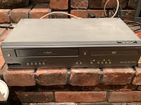 Front view of the Magnavox DVD/VCR combo player placed on a brick surface, showing the DVD tray, VCR slot, buttons, and indicator lights on the front panel.