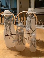 Group photo of the four frosted glass snowmen on a wooden table in front of wooden chairs. Visual confirmation of size and style, showing frosted glass bodies, clear heads, glitter accents, and silver twig arms.