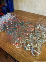 Thirty holiday-themed drinking glasses arranged on a table showing several rows of glasses with colorful village winter scenes and candy cane motifs.