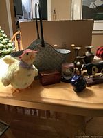 Overview of all items on wooden table including singing chicken, metal wood bin, wooden card box, pottery bunny, brass vases, rolling cow, and coasters