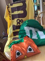 Three kids Halloween costumes: yellow M&M's bag, green M&M, and orange pumpkin costume on a chair.