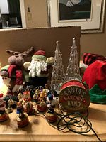 Overall view of lot including Santa's Marching Band ornaments, Santa and reindeer plush shelf sitters, silver metal tree tea light holders, and red-green singing hat.
