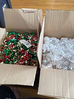 Open cardboard boxes showing large amounts of peel and stick curly ribbon bows in two colors: one box with white bows and the other with multi-coloured red, green, and gold bows.