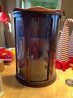 Front view of small glass and wood china cabinet showing curved glass front door with metal latch handle, wood top with curved trim, and dual glass side panels.
