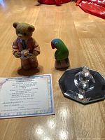 Overview showing Cherished Teddies Rick bear figurine holding a white animal, hand carved painted wooden parrot, and adoption certificate on wooden surface.