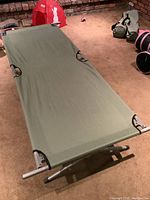 Full view of one green canvas camp cot showing stretched fabric over metal frame on a carpeted floor.