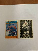 Photo showing two hockey cards placed side-by-side on paper: on the left, the 1982-83 OPC hockey card featuring Mark Messier and Wayne Gretzky in Edmonton Oilers uniforms, showing a crease; on the right, the Topps Marcque Men Wayne Gretzky card in black and white with gold border.