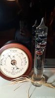 Round wooden-cased barometer and Galileo thermometer set shown with reflections in background.